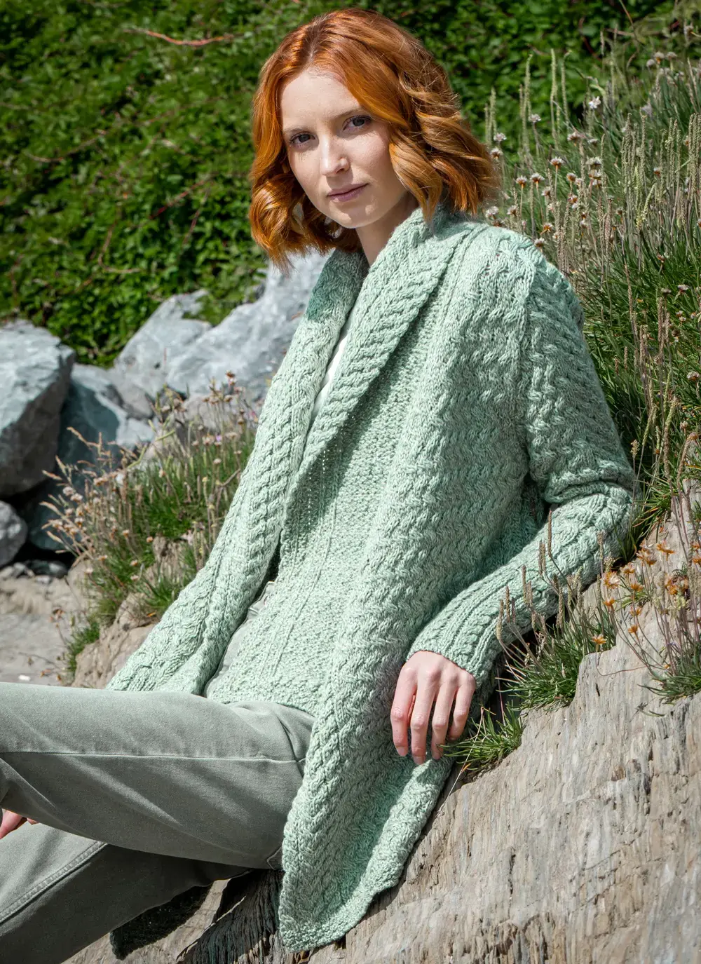 Red headed model sitting on rock in front of greenery in sage green glenross aran waterfall cardigan