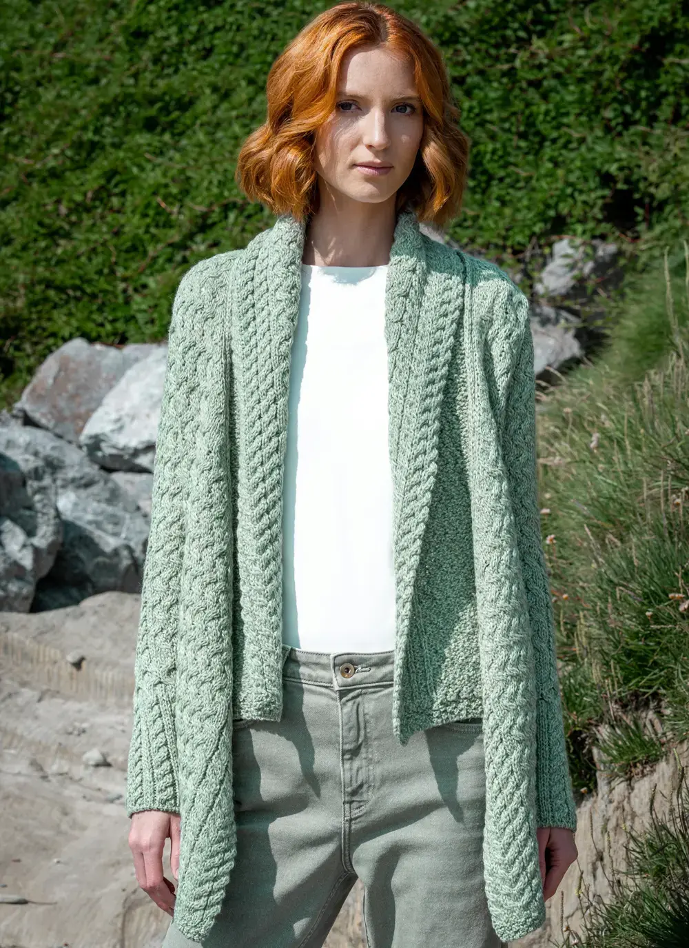 Red headed model standing in front of greenery in sage green glenross aran waterfall cardigan