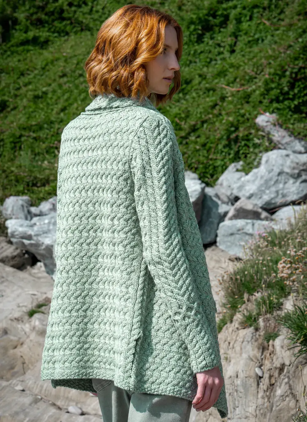 Red headed model turned looking over shoulder in front of greenery background in sage green glenross aran waterfall cardigan