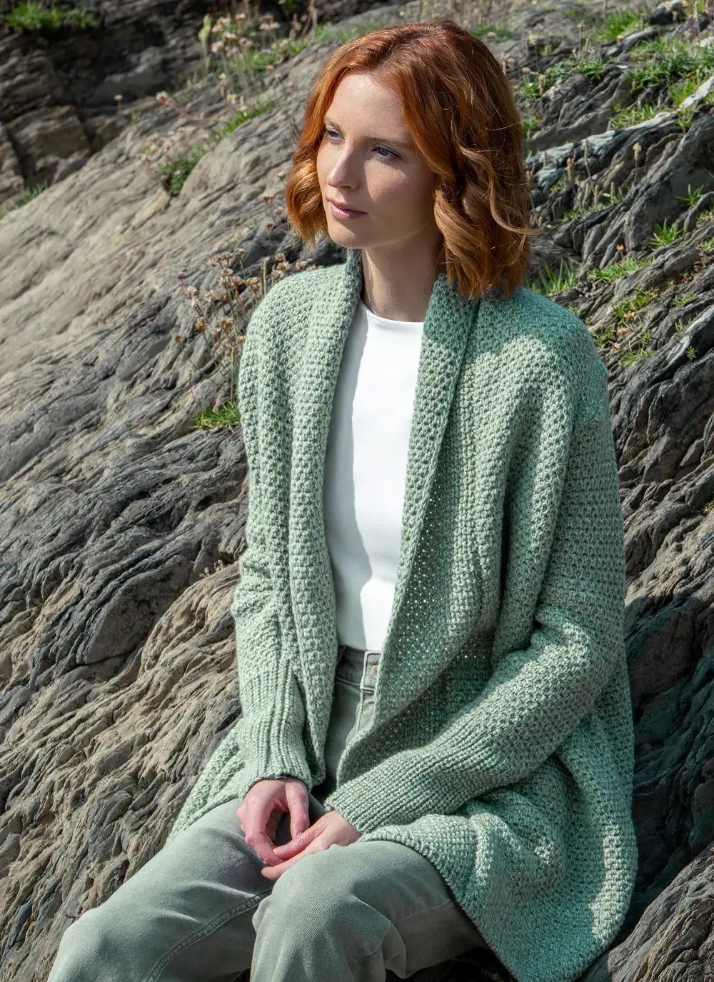 Red headed model sitting on rock wearing a sage green knitted kilcoole textured coatigan