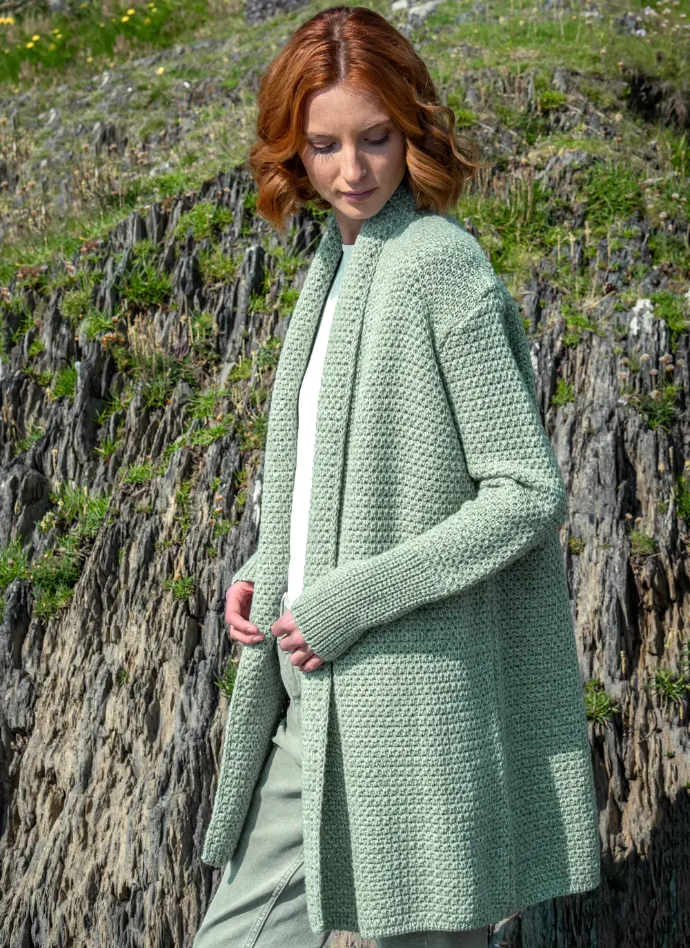 Red headed model standing sideways in rocky area wearing a sage green knitted kilcoole textured coatigan