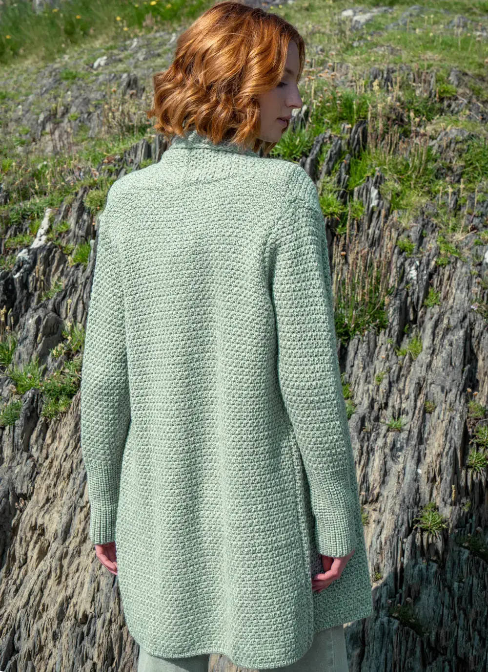 Red headed model facing back in rocky area wearing a sage green knitted kilcoole textured coatigan