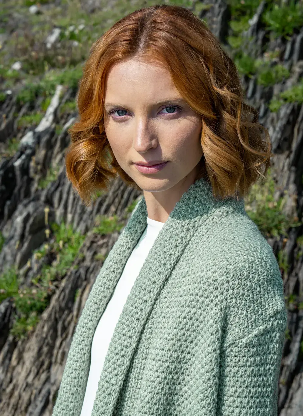 Red headed model detail shot of collar in front of rocky area wearing a sage green coatigan