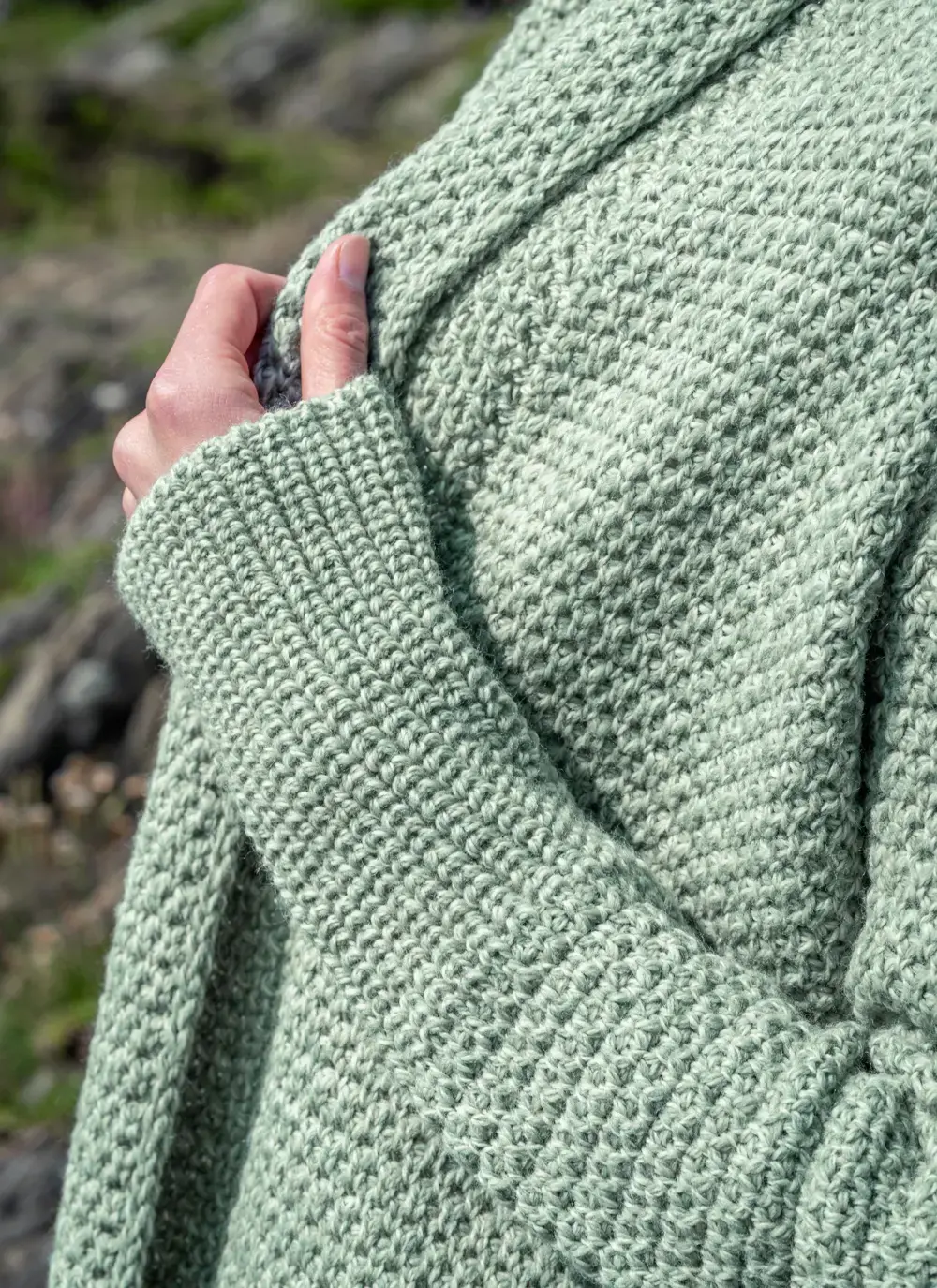Red headed model detail shot of sleeve in front of rocky area wearing a sage green coatigan