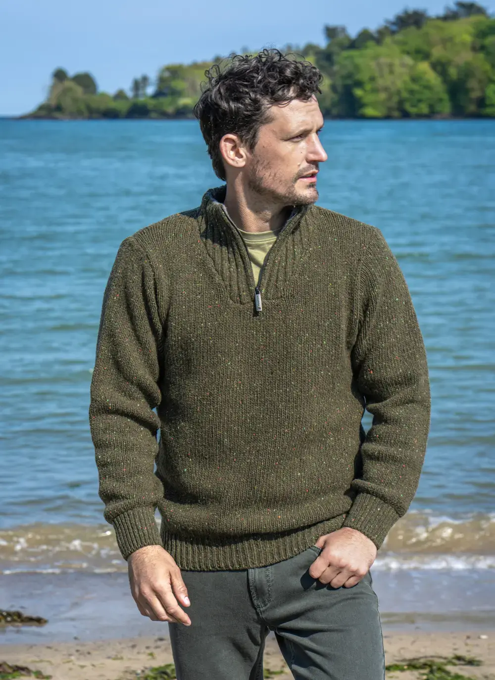 Man wearing Glenstone Contrast 1/2 Zip Sweater in green flecked wool, standing by the shoreline with blue sea in the background.