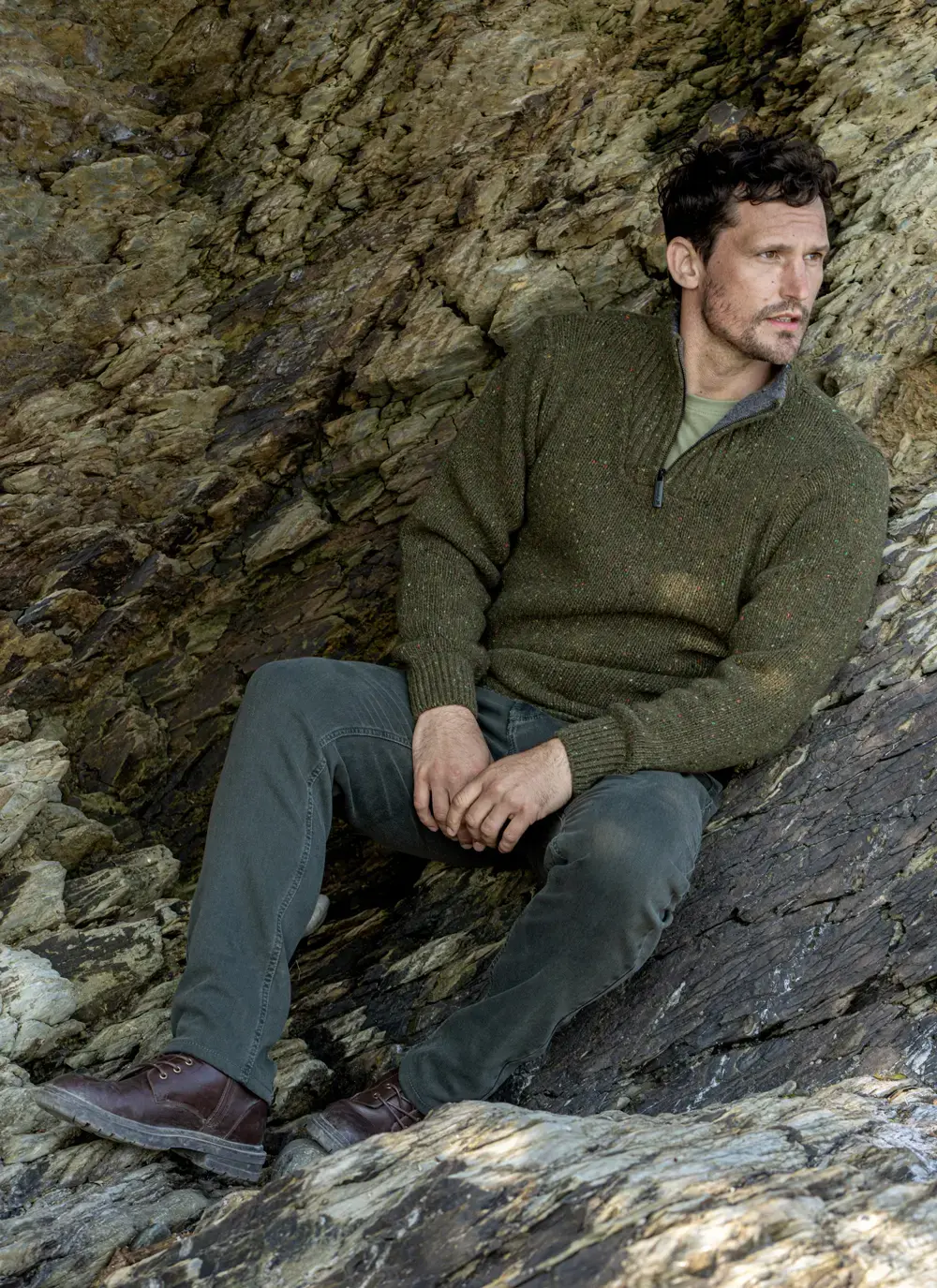 Model seated against natural rock face wearing Glenstone Contrast Zip Sweater and dark green jeans with leather boots.