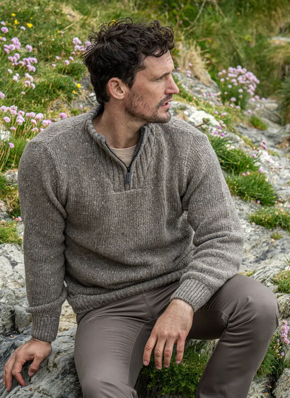 Model sitting among wildflowers and rocks, wearing Glenstone Contrast 1/2 Zip Sweater in Rocky Ground with taupe trousers.