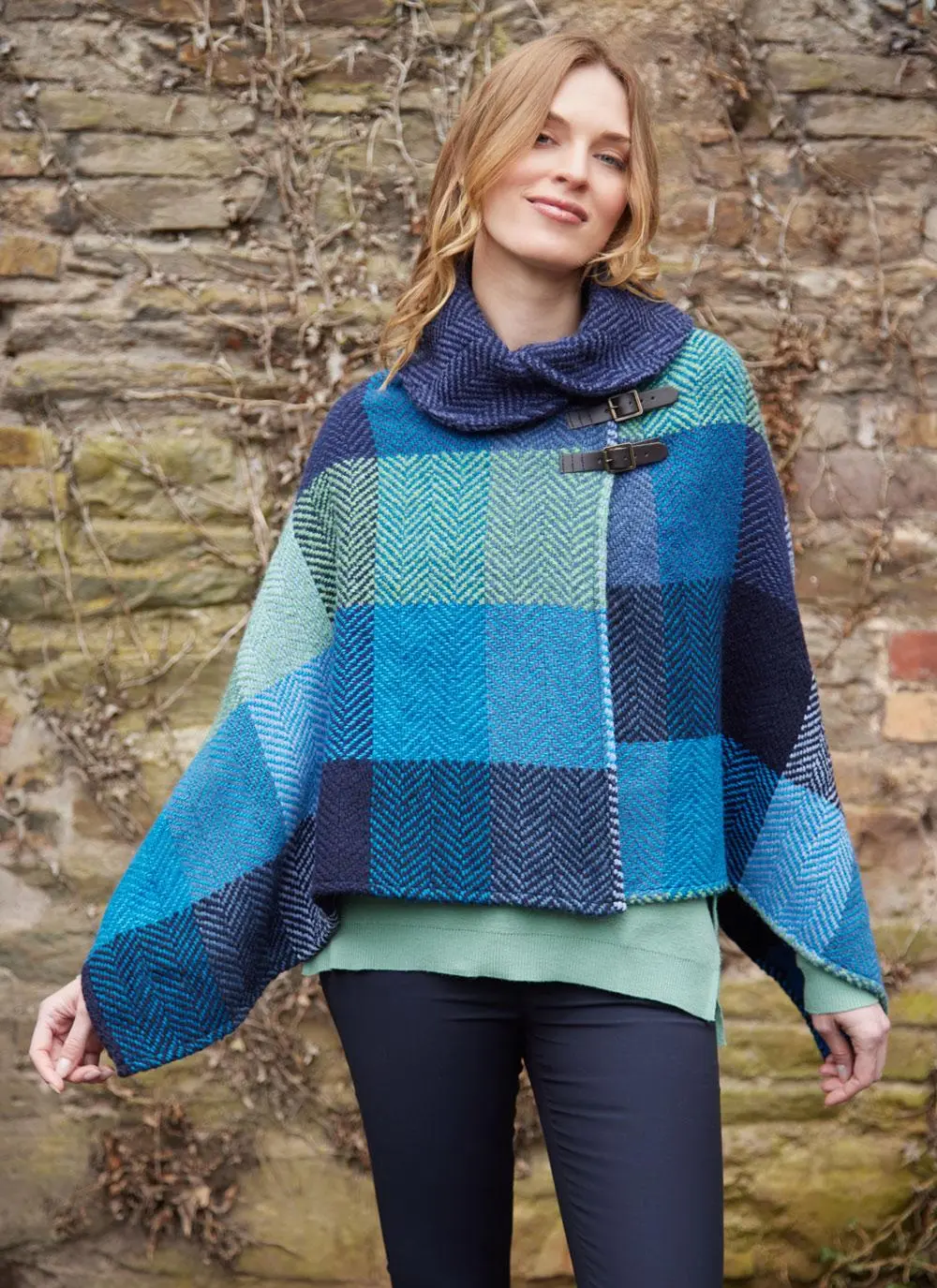 Blonde smiling woman wearing jeans and blue check poncho with shawl buckle collar against brick wall backdrop.