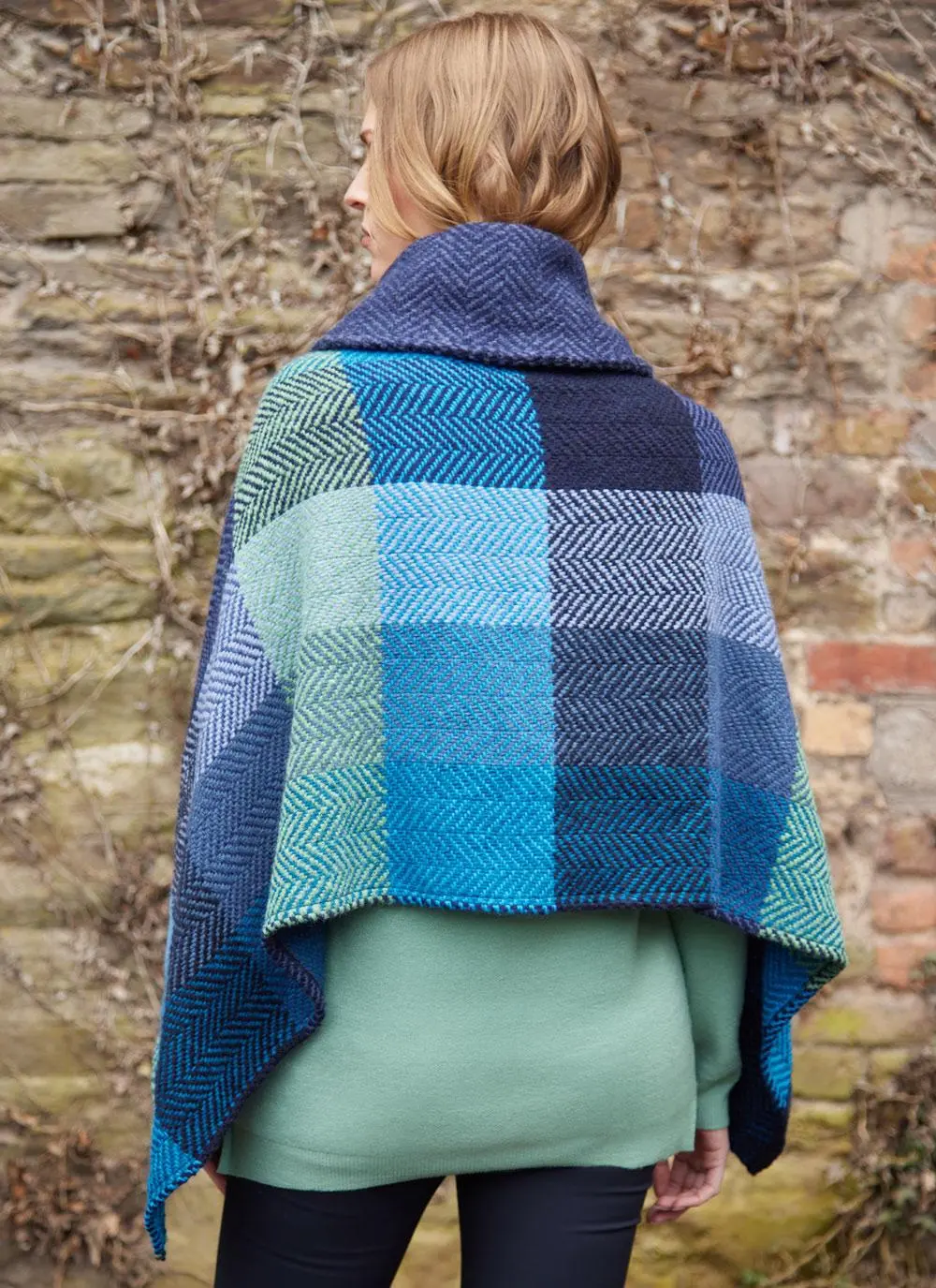 Back angle shot of blonde woman wearing jeans and blue check poncho with shawl buckle collar against brick wall backdrop.