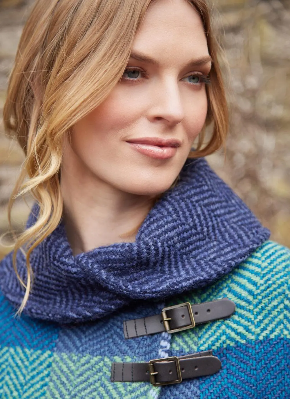 Close up shot of blonde woman wearing blue check poncho with shawl buckle collar against brick wall backdrop.