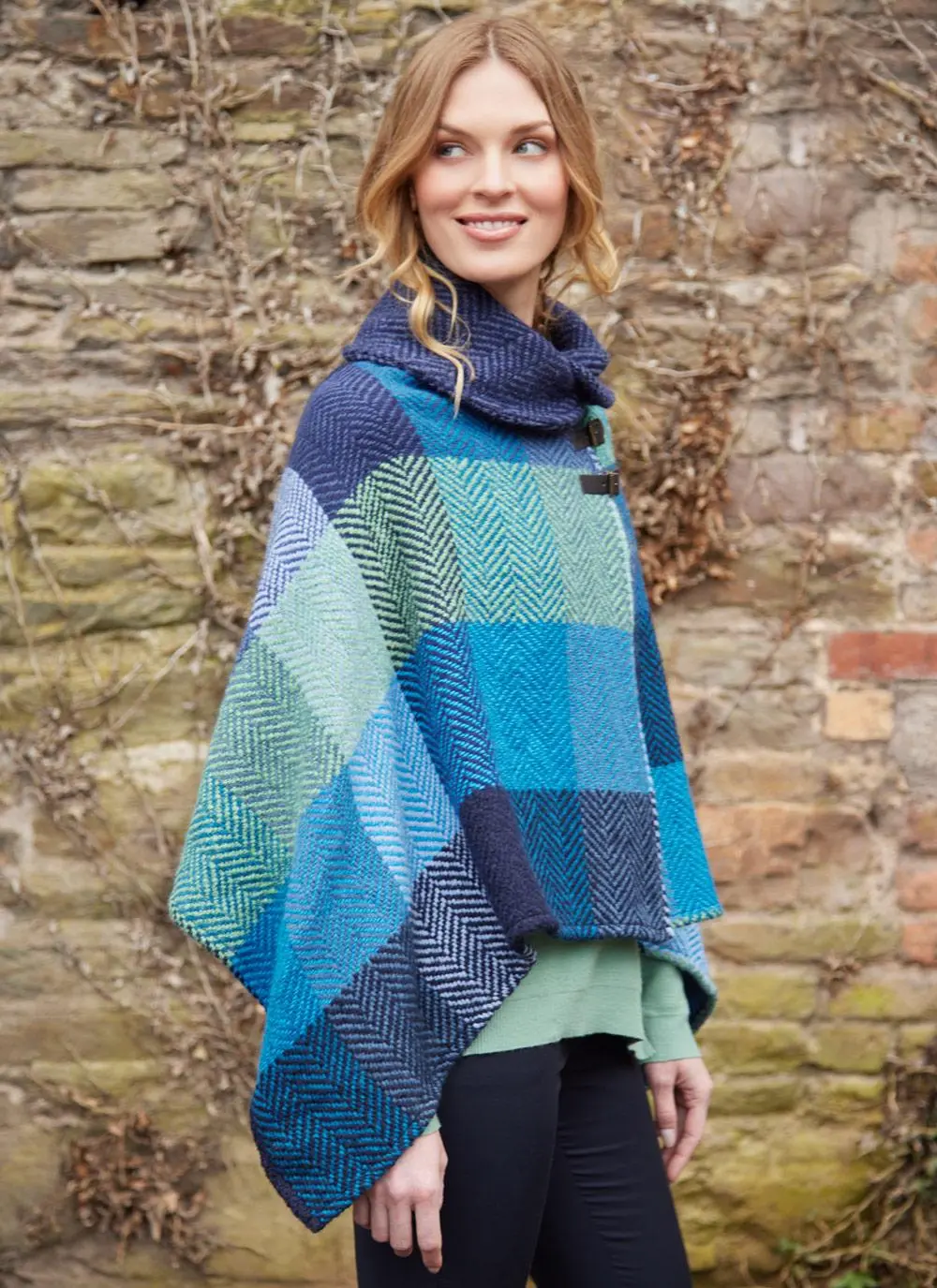 Blonde woman wearing jeans and blue check poncho with shawl buckle collar against brick wall backdrop.