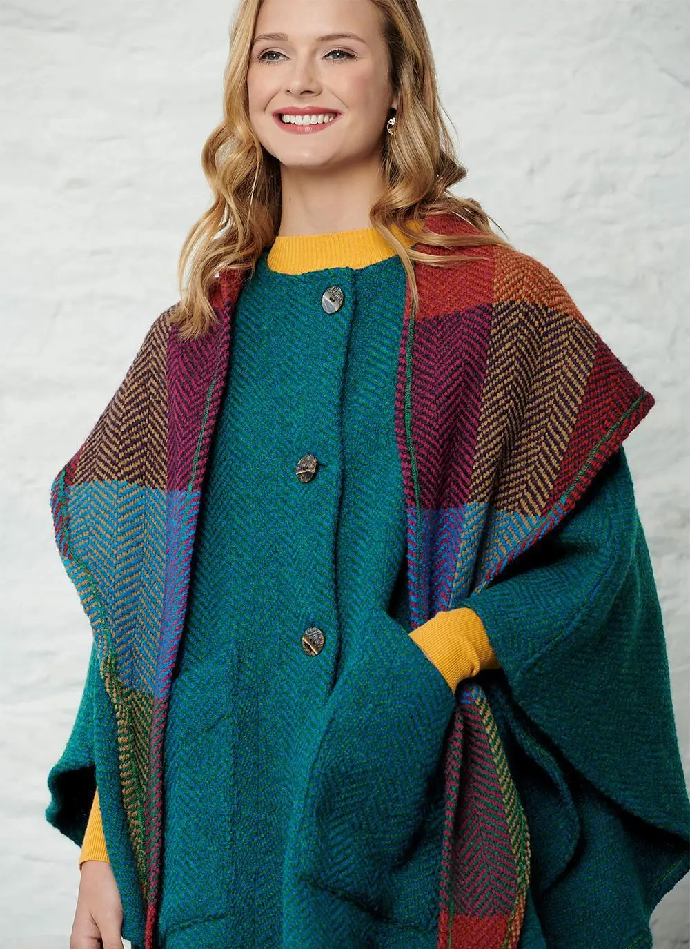 Close up studio shot of women wearing green cape with detachable multi-colored hood with both hands in pockets.