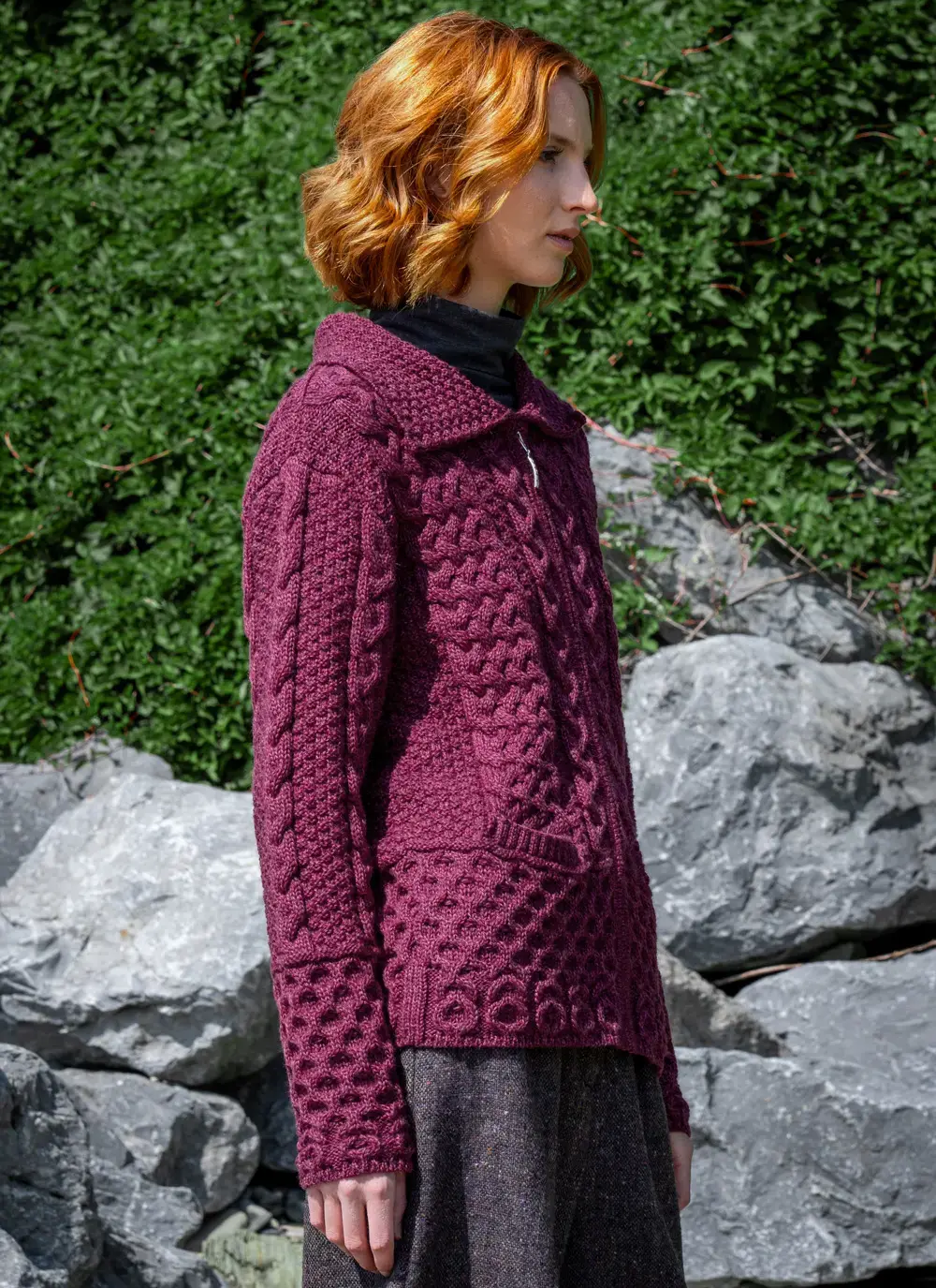 Red head model sideways in front of greenery in grace full zip aran cardigan in raspberry melange