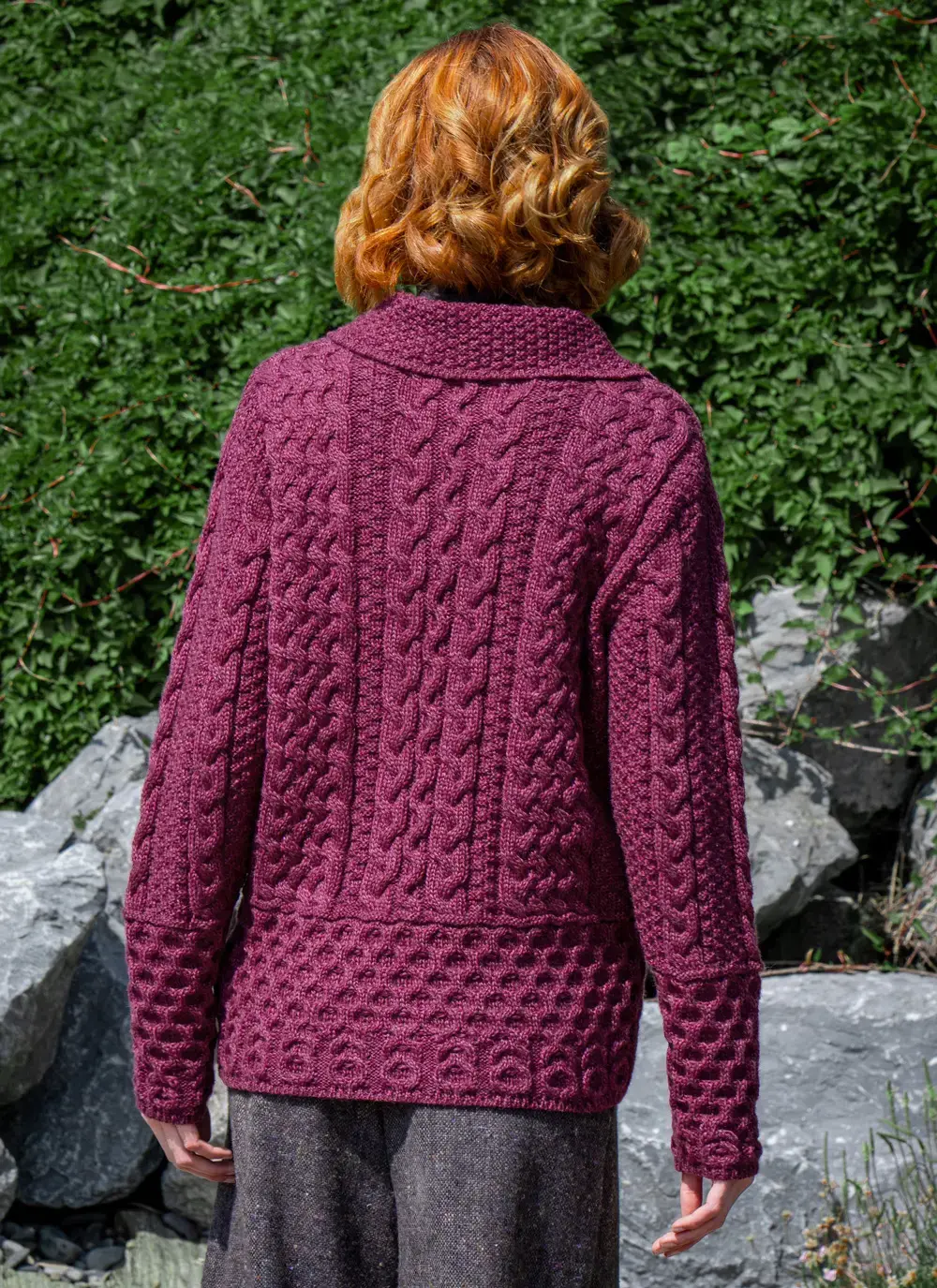 Red head model backwards in front of greenery in grace full zip aran cardigan in raspberry melange
