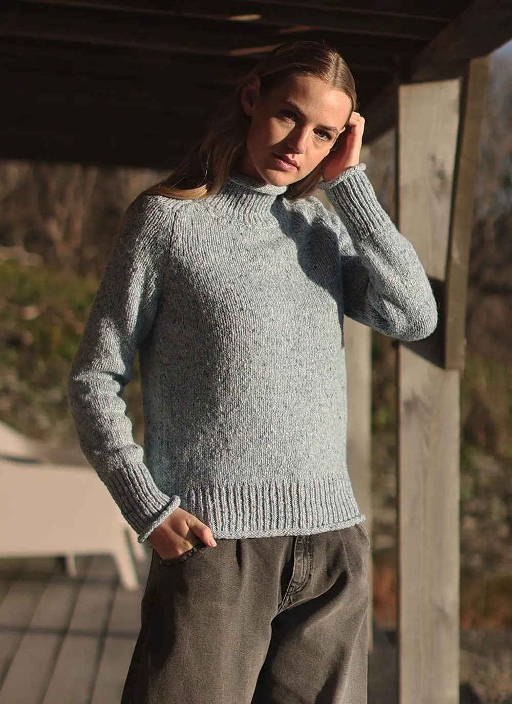 blonde woman wearing blue sweater with stable in background, one hand in pocket one touching face