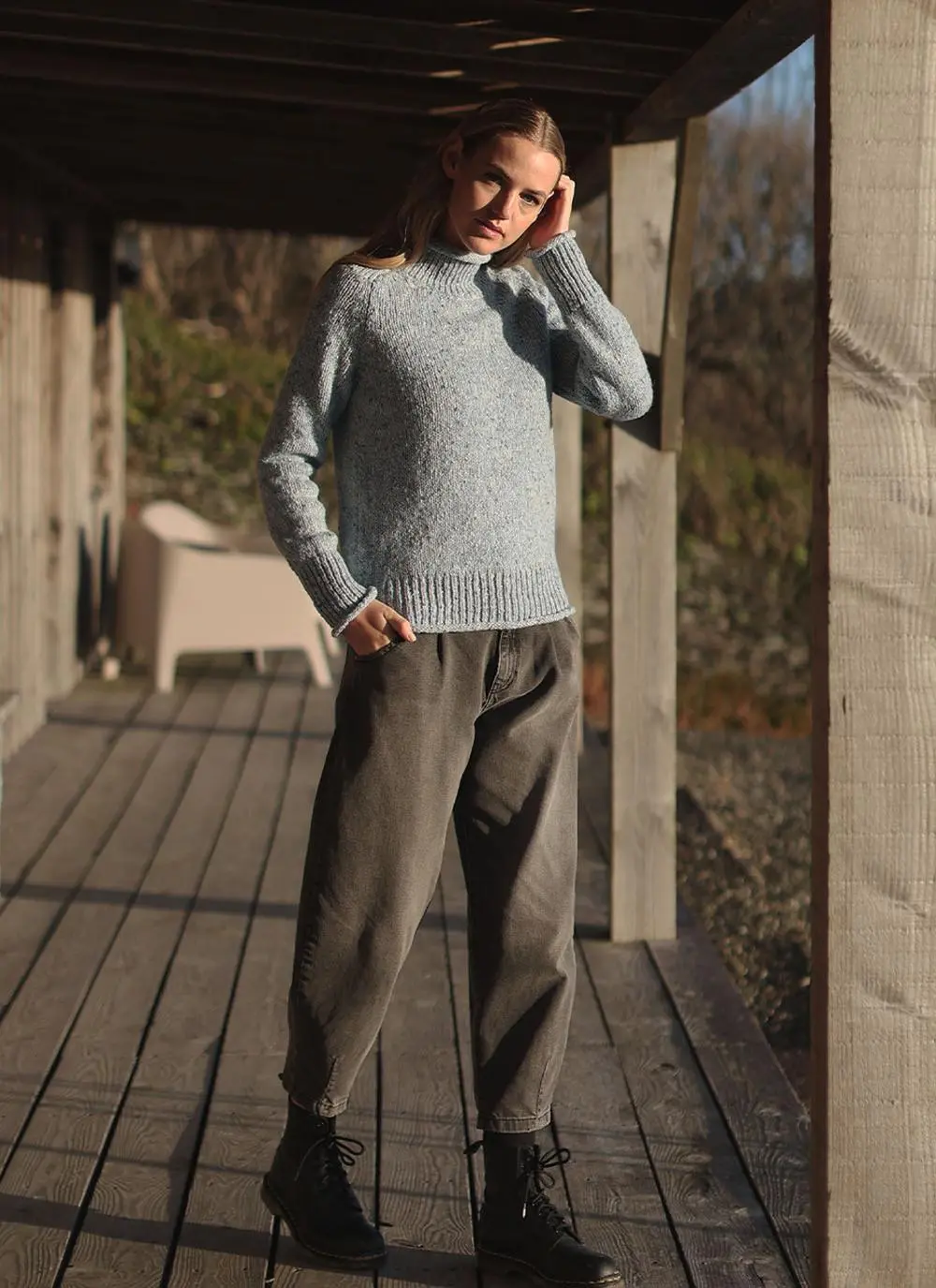 full body shot of blonde woman wearing blue sweater with stable in background