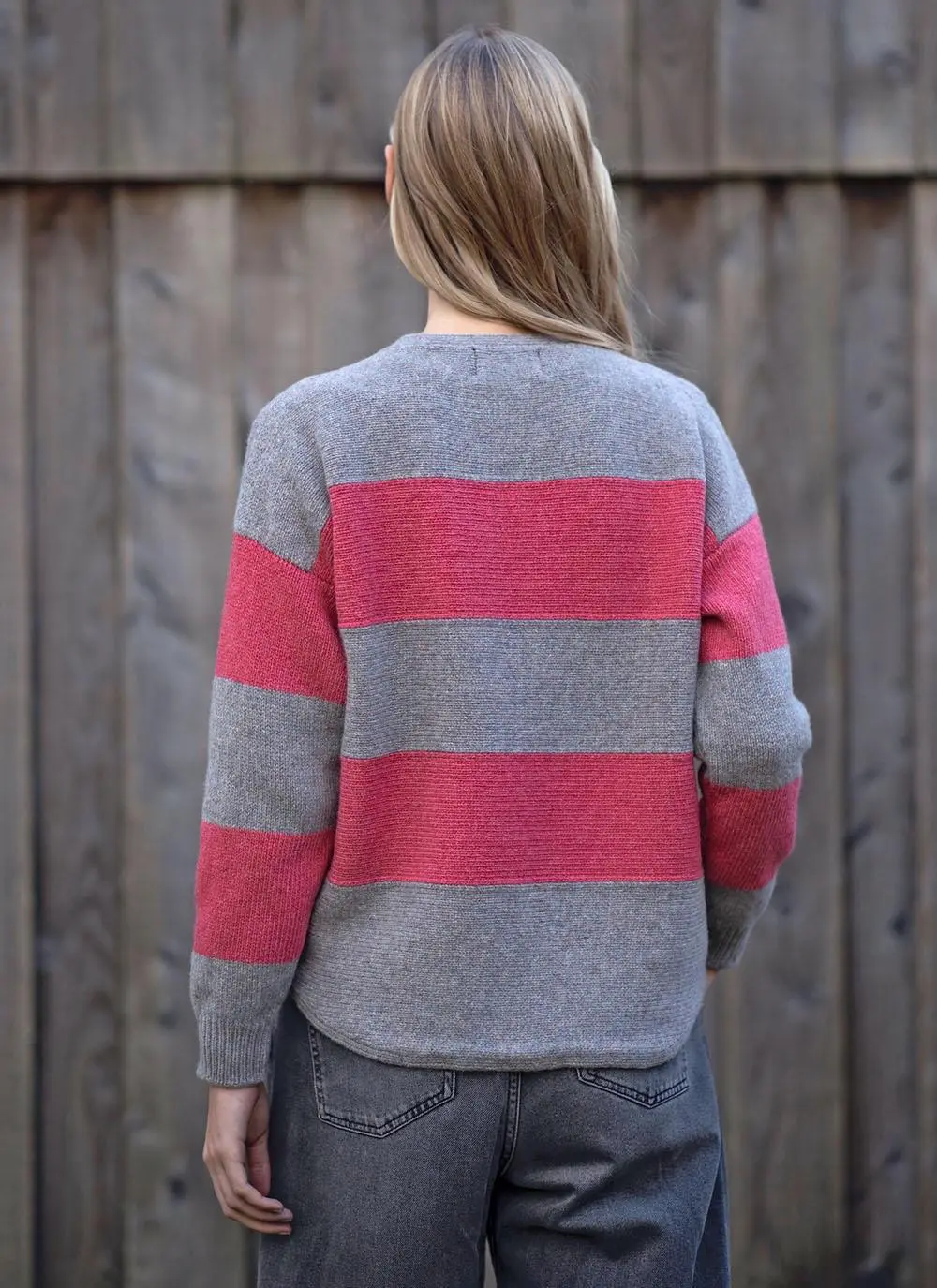 back shot of blonde woman standing in front of wooden wall wearing grey and pink stripe sweater