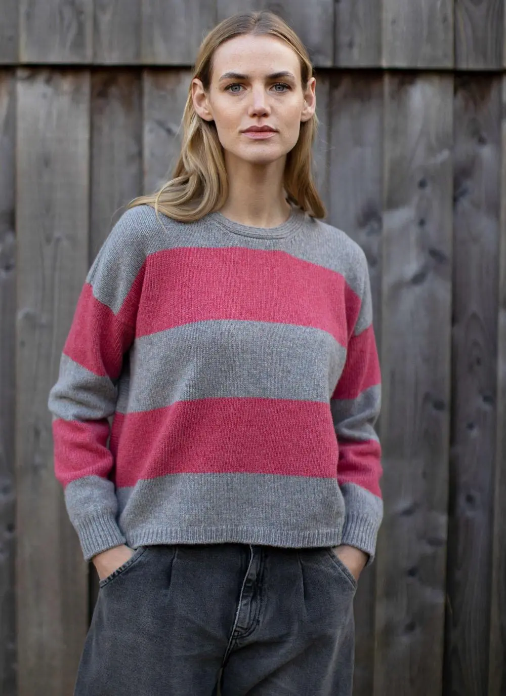 blonde woman standing in front of wooden wall wearing grey and pink stripe sweater with hands in pockets