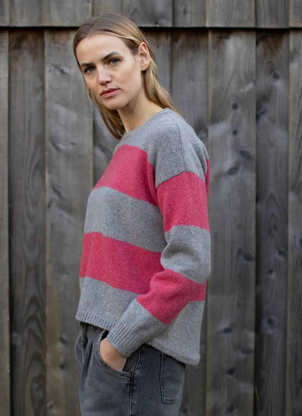 side angle of blonde woman standing in front of wooden wall wearing grey and pink stripe sweater with hands in pockets