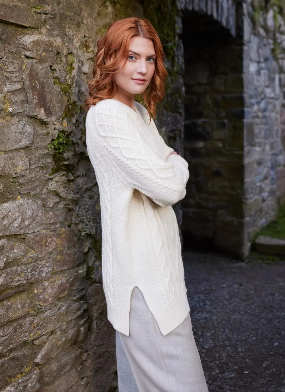 Side shot of red haired woman in abbey wearing white Aran sweater with both arms folded