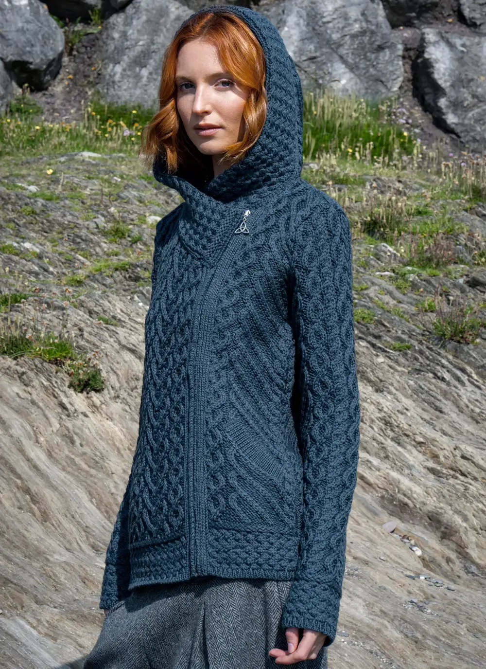 Red headed model in rocky area wearing a blue Aran biker hoodie standing to the side with hood up
