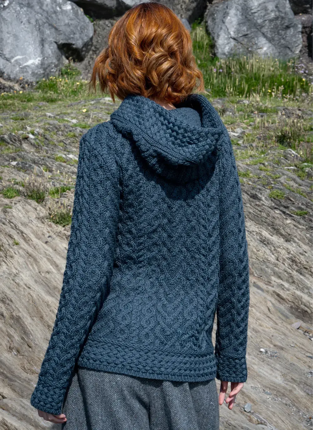 Red headed model in rocky area wearing a blue Aran biker hoodie back view