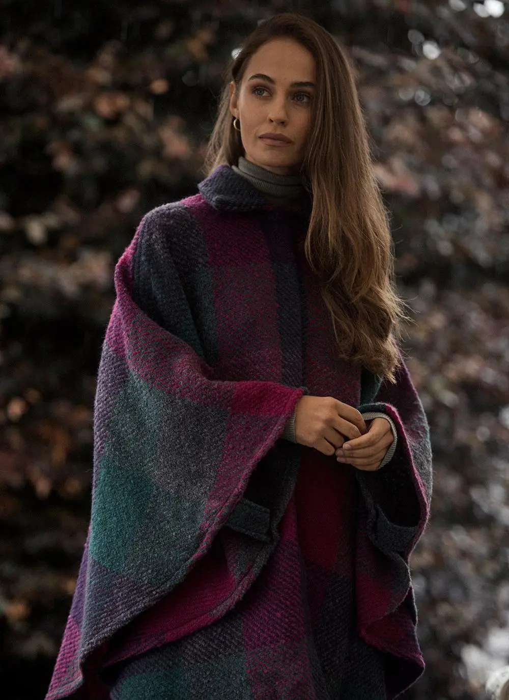 Woman standing against stony wall wearing wool purple toned cape with hands clasped.