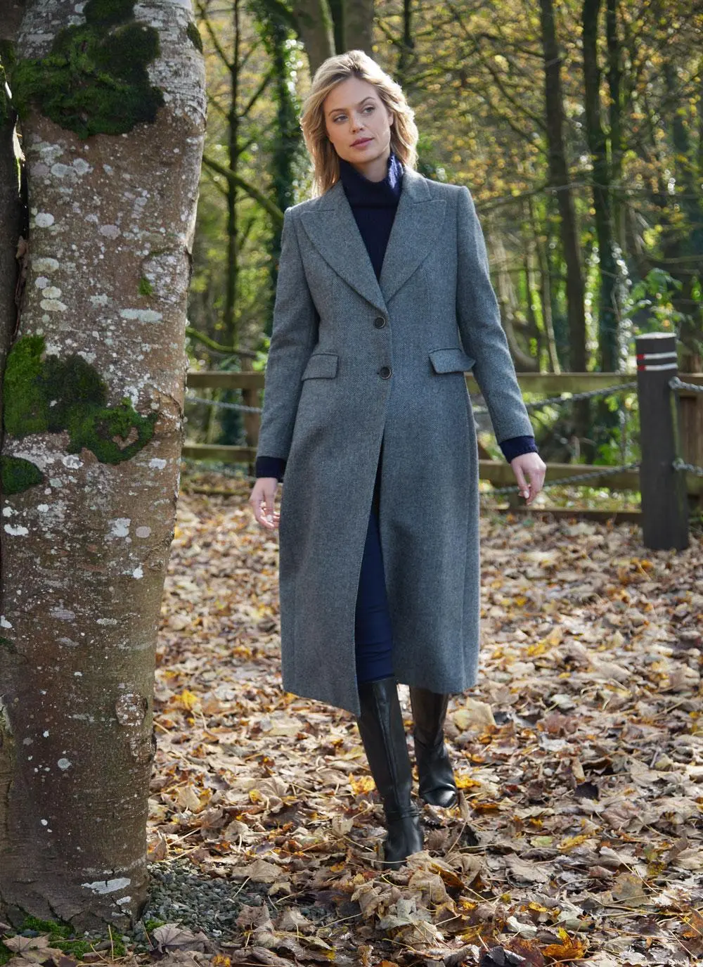 Blonde woman walking forward in woodland area wearing longline blue herringbone tweed coat