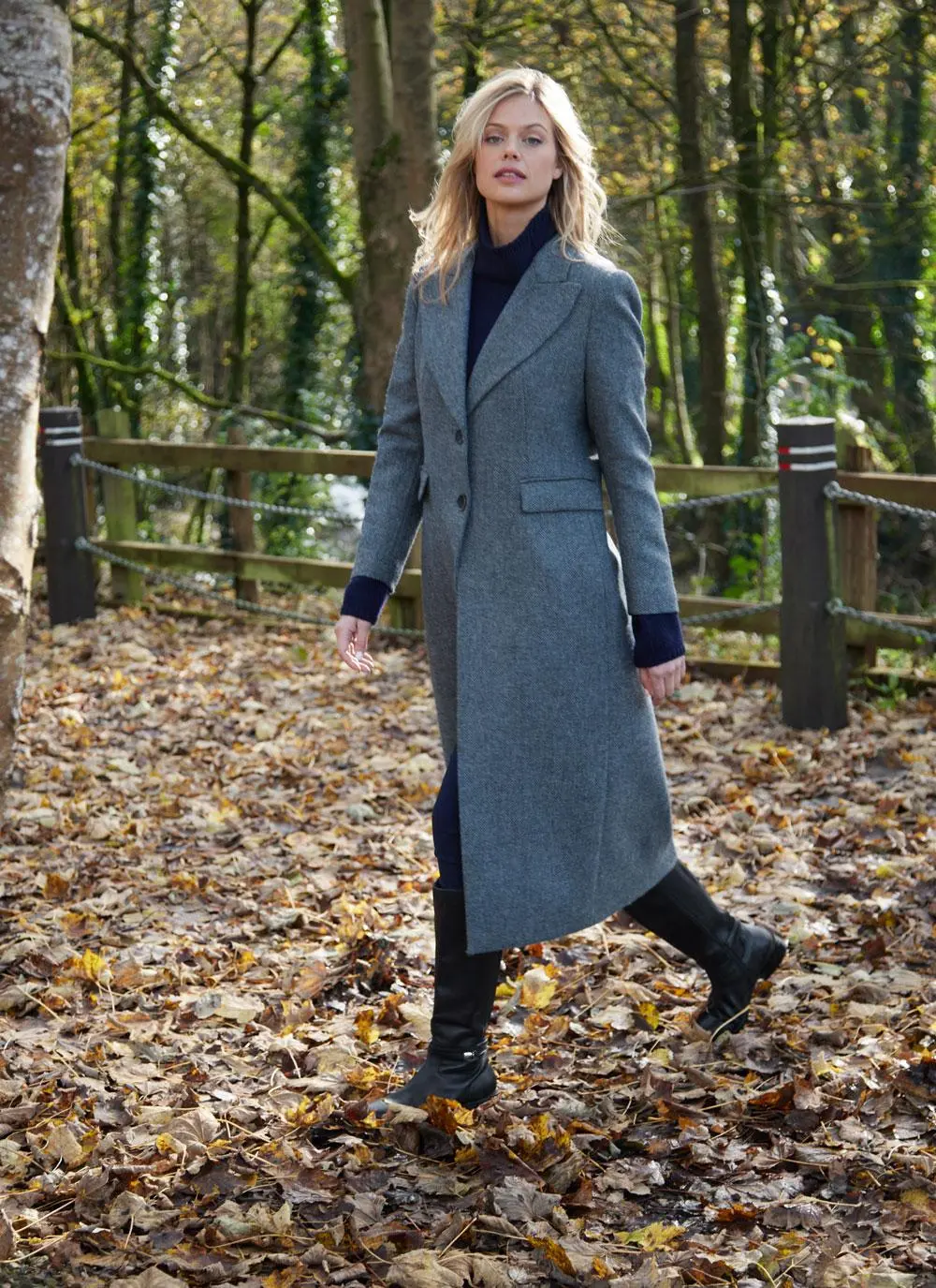 Blonde woman walking in woodland area wearing longline blue herringbone tweed coat