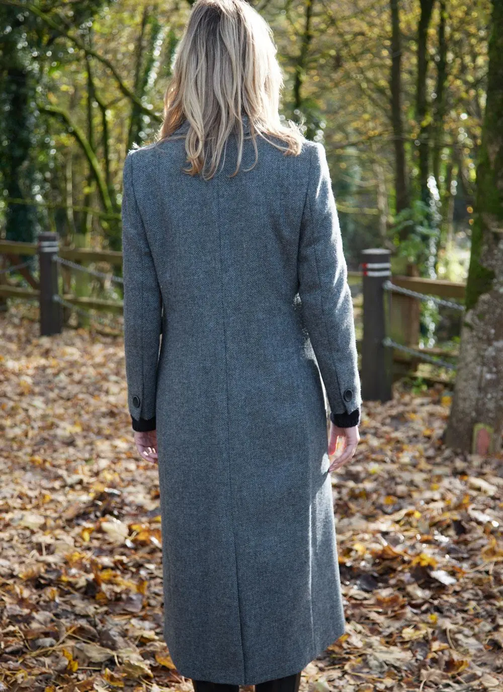 Back shot of blonde woman in woodland area wearing longline blue herringbone tweed coat