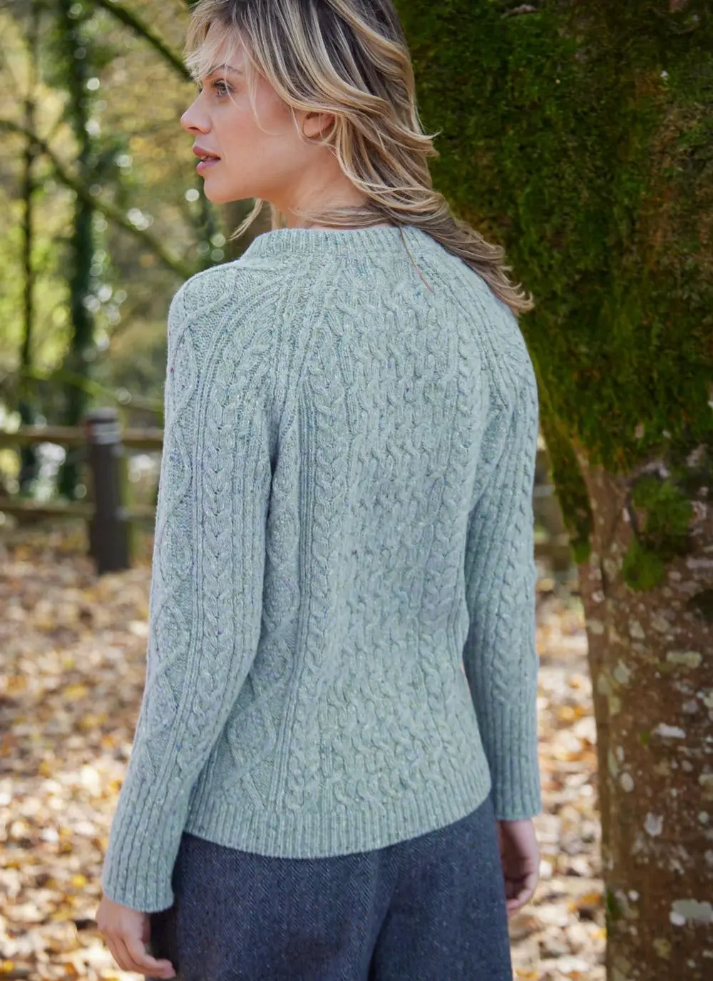 A woman in a green Aran Wool sweater and gray trousers stands in a wooded area, with autumn leaves and trees in the background.