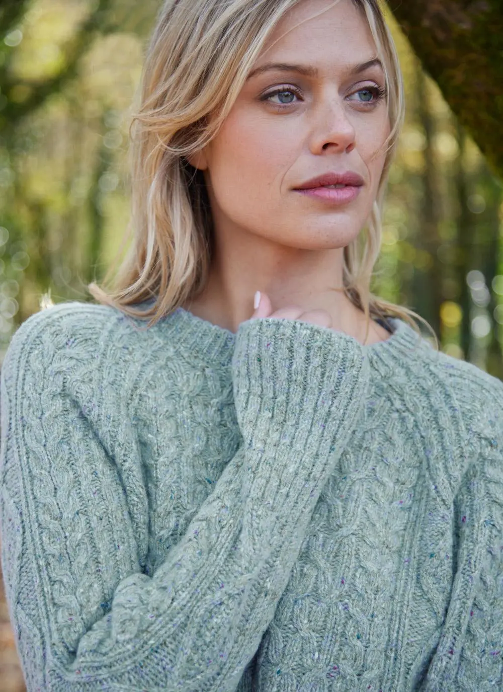 A woman in a green Aran wool sweater stands in a wooded area, with green trees in the background.