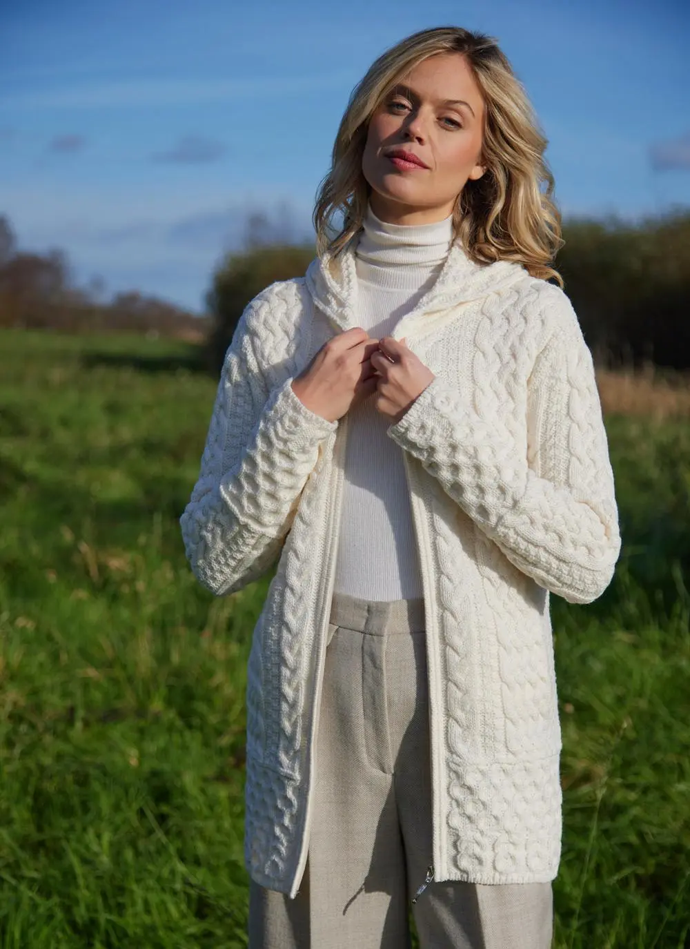 A blonde woman standing in field wearing celtic aran natural hoody zipped down