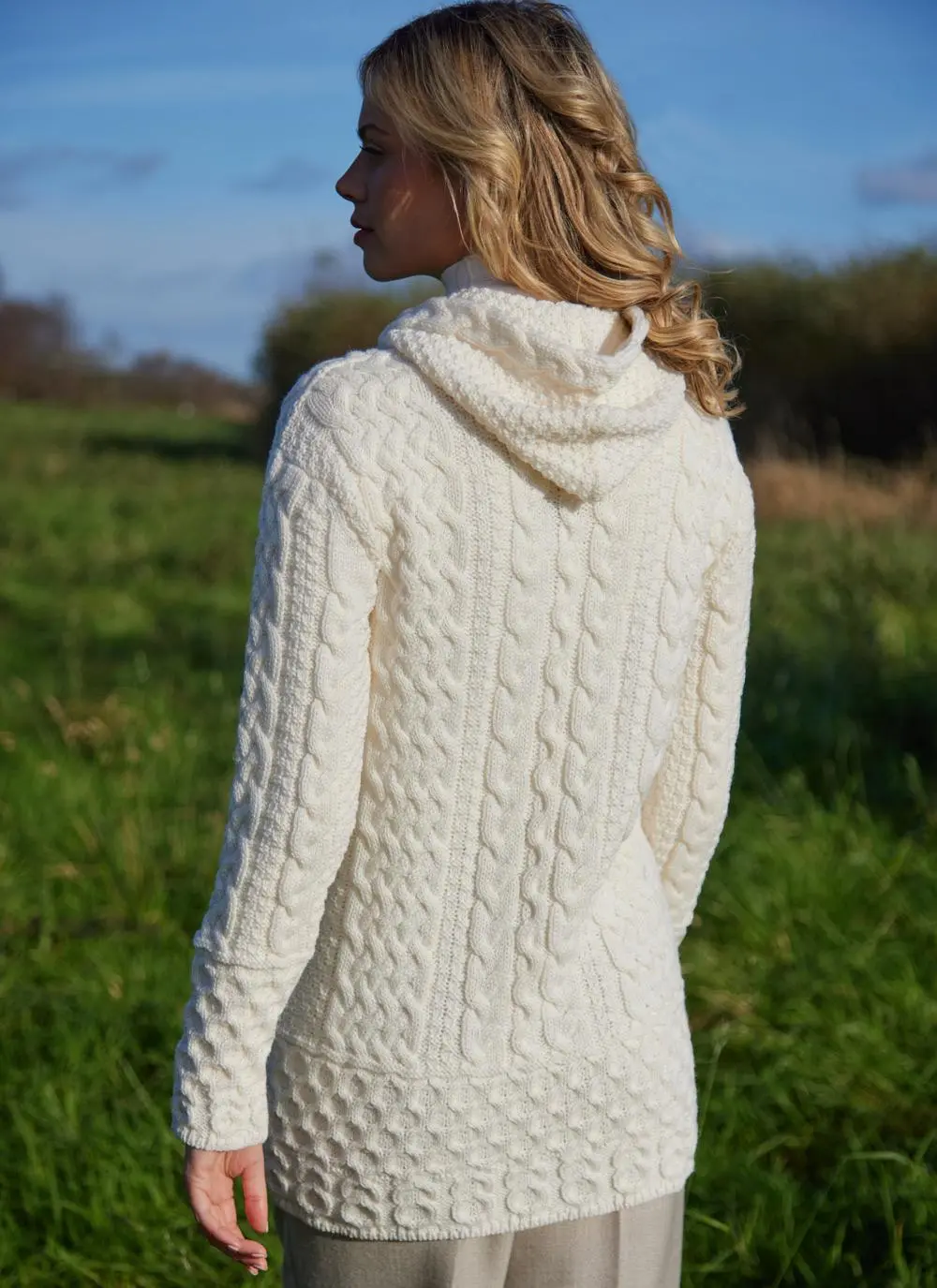 A blonde woman standing in field wearing celtic aran natural hoody, back shot