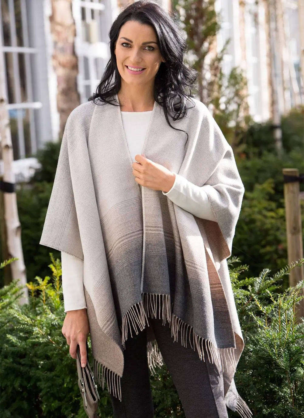 Brunette woman wearing black and beige fringed shawl with building and greenery in background. 