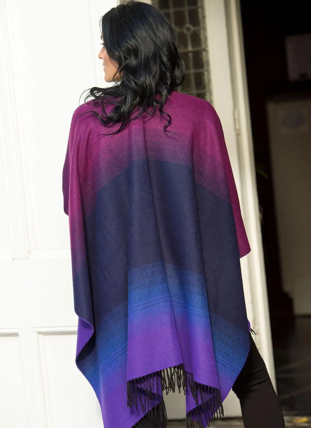 Back angle shot of brunette woman wearing purple and black shawl standing in front of building. 