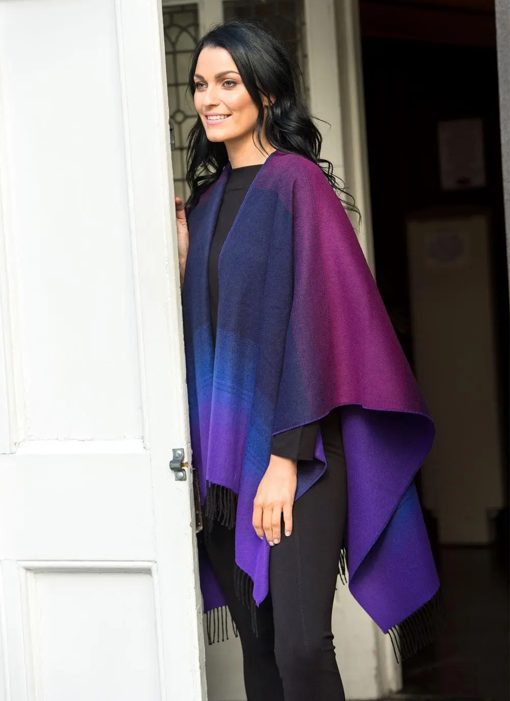 Brunette woman wearing purple and black shawl standing in front of building. 