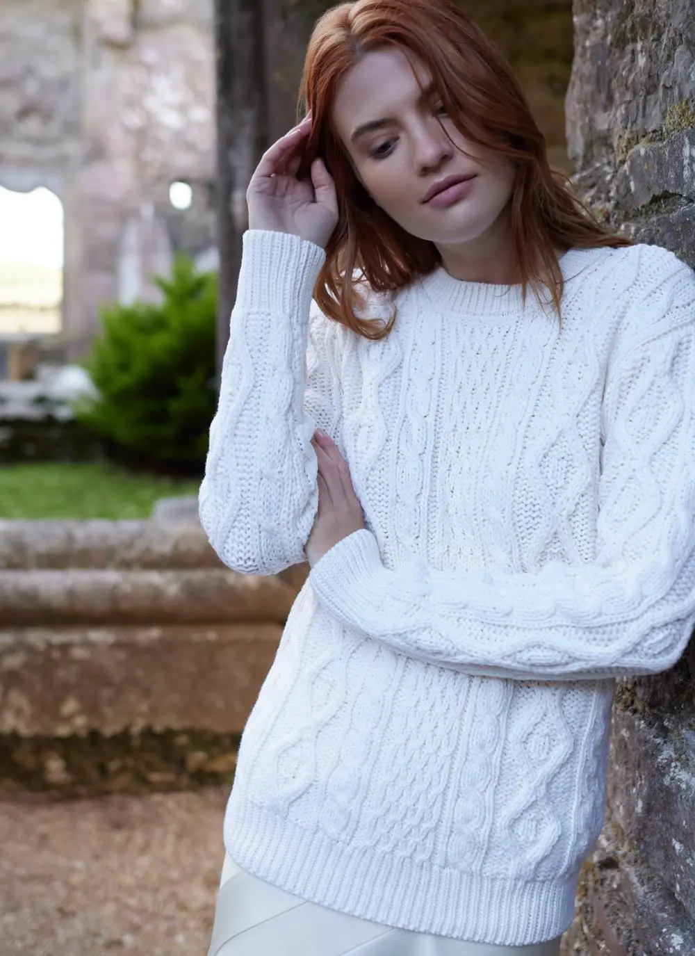 red haired woman leaning against wall in stony passageway with arms folded wearing a white aran sweater
