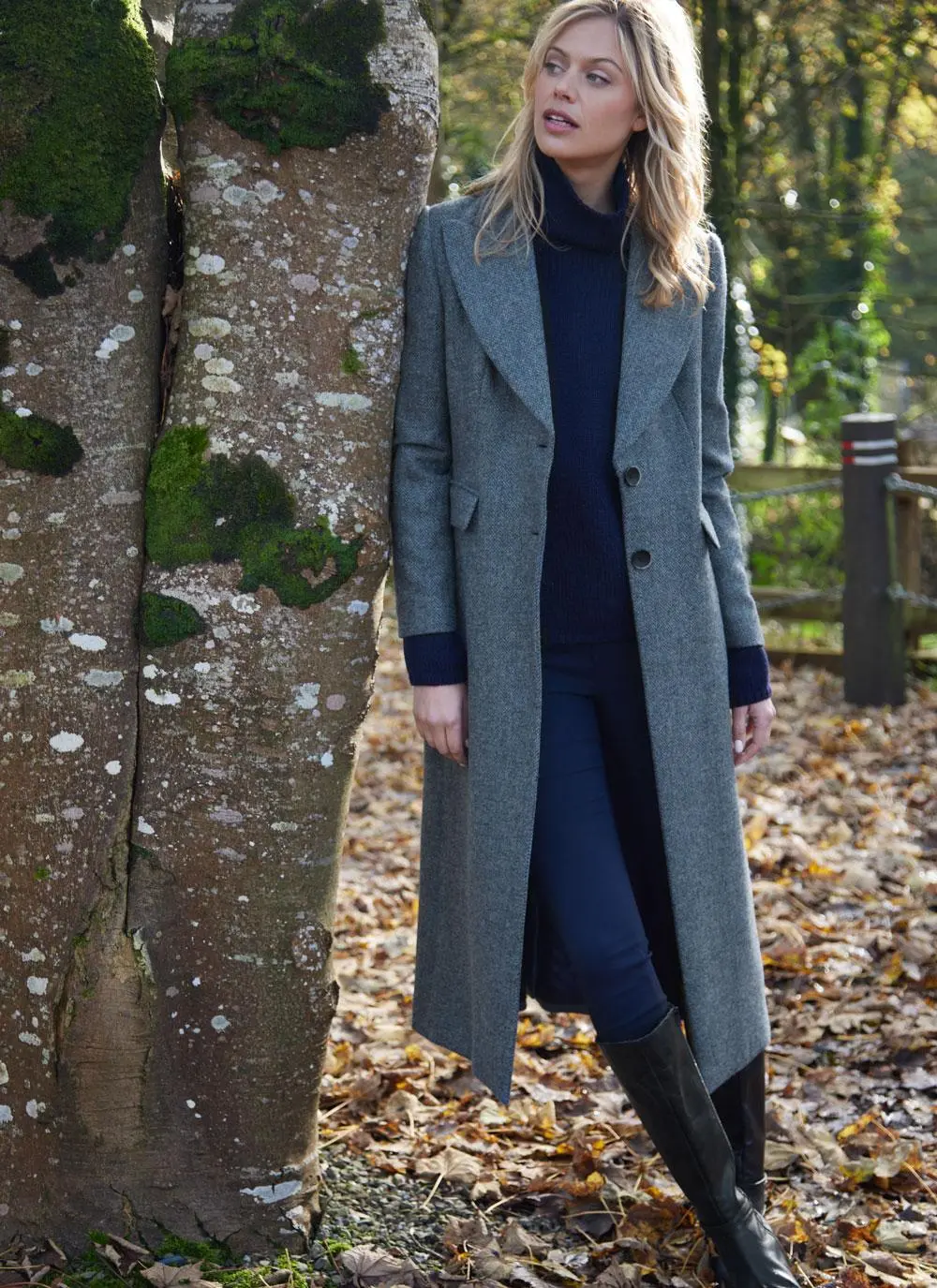 Blonde woman in woodland area wearing longline blue herringbone tweed coat with buttons open