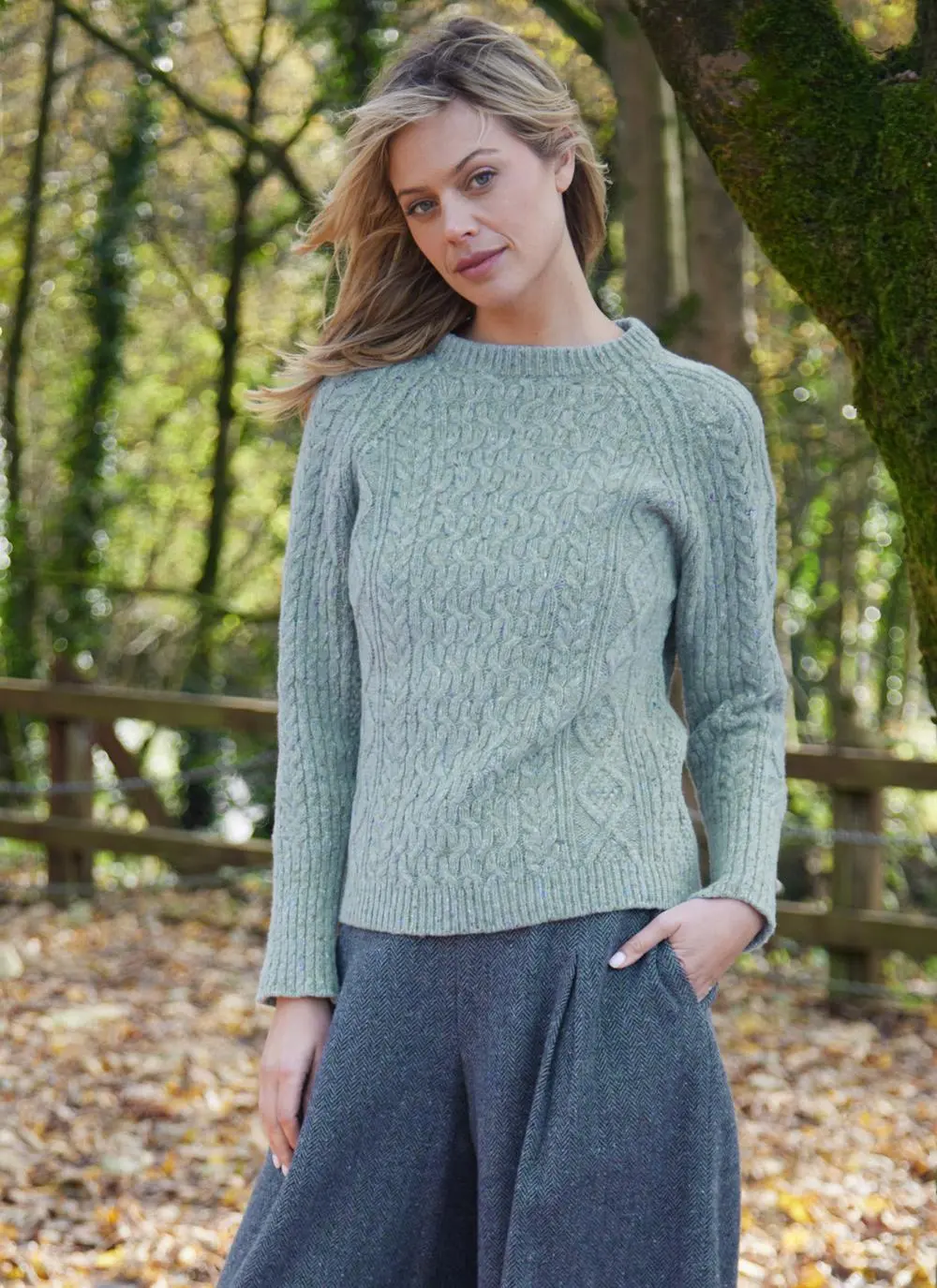 A woman in a green Aran sweater and gray trousers stands in a wooded area, with autumn leaves in the background.
