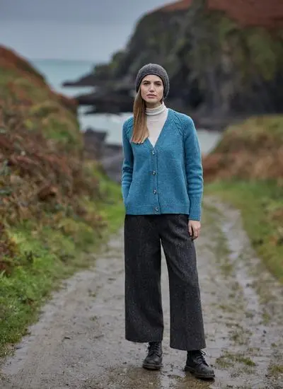 woman standing in path in grassy outdoor area wearing blue cardigan, tweed pants and a grey beanie