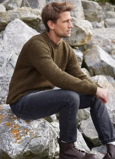 Man sitting on a rock on a beach wearing an olive flecked fisherman crew neck sweater