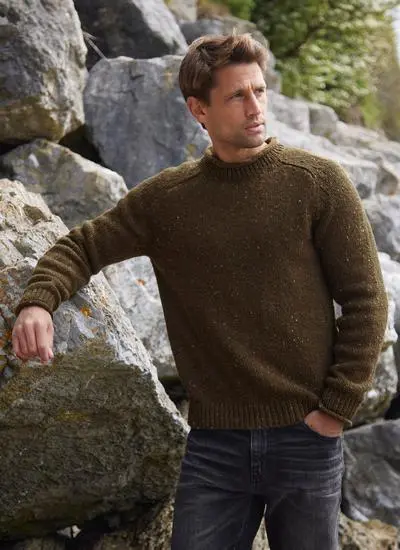 man leaning on rocks with one arm wearing olive colored knit sweater, other arm in pocket