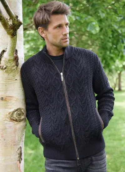 Man leaning on a tree wearing a black aran full zip cardigan, brown trimming on over zip and pocket