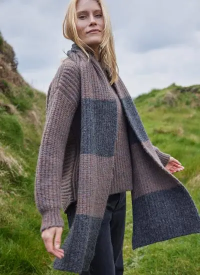 blonde woman standing in grassy coastal area wearing a copper and grey knit scarf blowing in wind