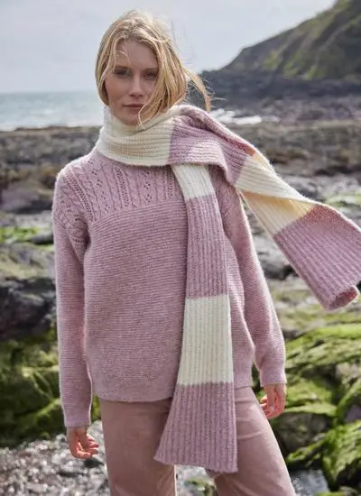 blonde woman standing on rocky, mossy beach wearing large knit pink and white scarf blowing in wind