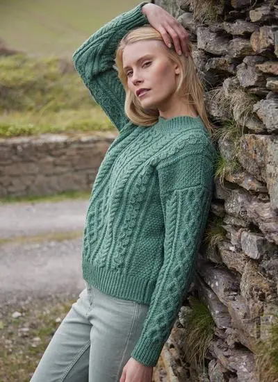blonde woman leaning against wall of stone cottage in grassy area wearing sage aran sweater