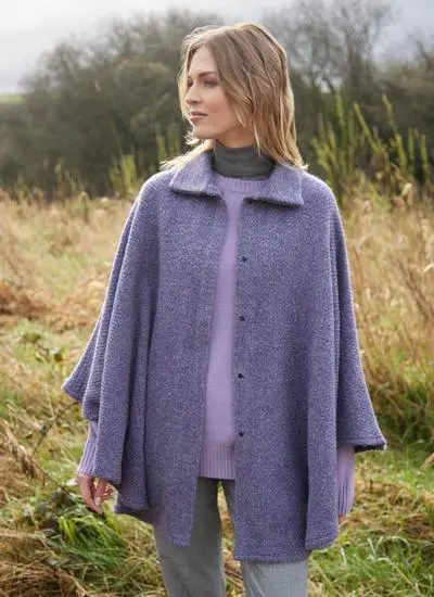 Blonde woman standing in Irish countryside wearing open lilac wool cape. 