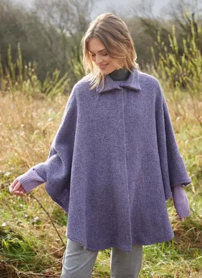 Blonde woman standing in Irish countryside wearing lilac wool cape. 