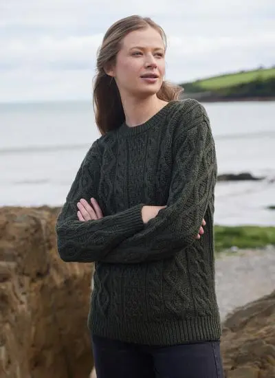 woman standing in stony beach wearing army green aran sweater with arms folded